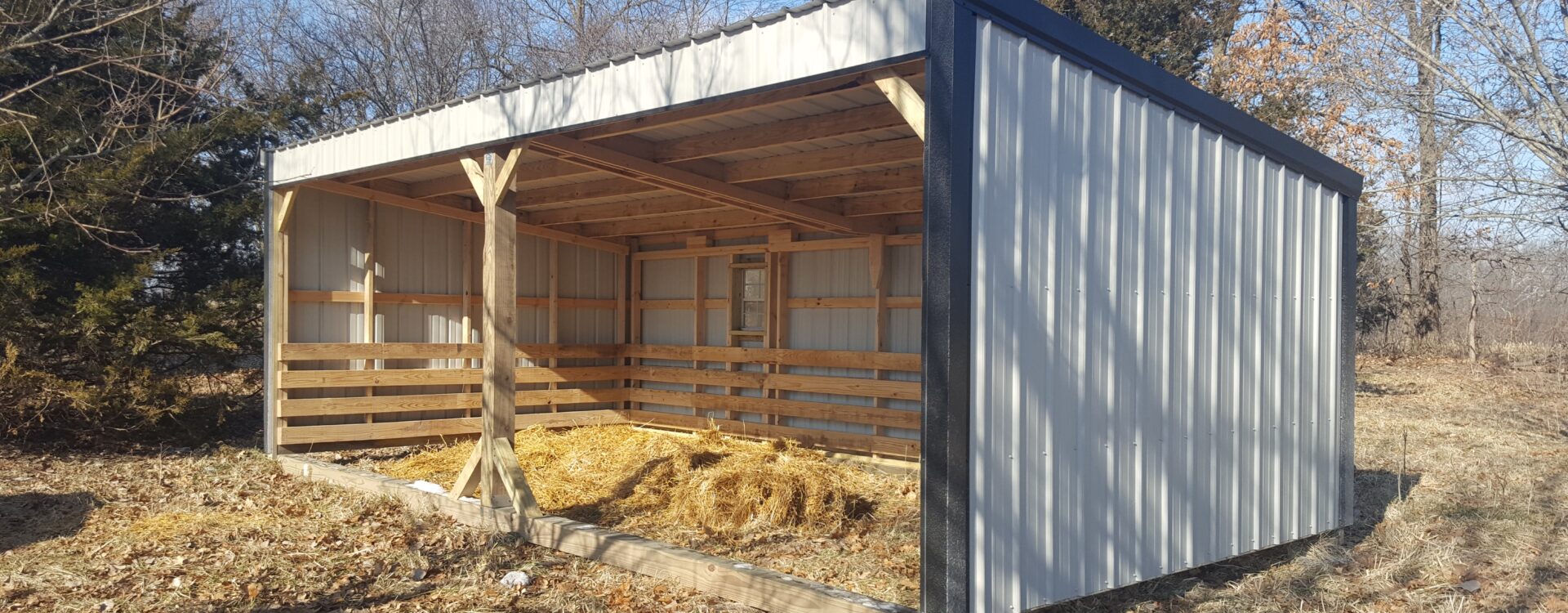 White and black metal loafing shed with hay on the ground - trees in background