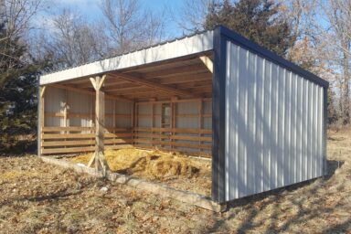 White and black metal loafing shed with hay on the ground - trees in background