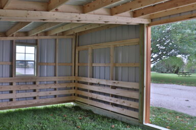 interior of metal loafing shed featuring wood beams supports and joists - blurred view out of side window and partial view of grassy picnic area beyond animal entrance