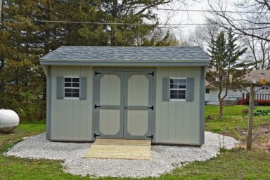 Kansas City Backyard Shed - Gray painted wood siding with medium gray-trimmed double doors corners and matching shutters on two windows beneath gray shingled roof - shed sits on gravel foundation in yard next to propane tank and evergreen trees - one-story white house with red deck visible nearby