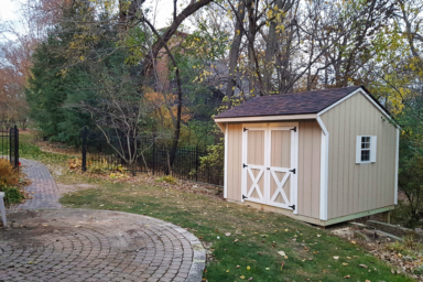 Des Moines Backyard Shed - Beige siding with white-trimmed double doors corners and side window with shutters - shed sits on edge of yard near black iron fence facing brick walkway