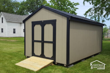 utility shed with ramp add on ia - Beige siding with black-trimmed double doors and corners beneath black metal roof - shed sits near tree with white two-story house nearby