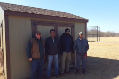 utility shed missouri - Tan siding with brown-trimmed double doors on shor and long sides - four men stand in front of shed facing camera