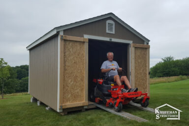 wooden shed in - taupe painted wood siding with white corner trim - smiling partially bald man descends ramps on his red and black lawnmower