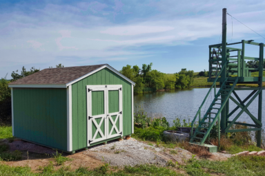 green utility shed ia - green painted wood siding with white trimmed double doors and corners beneath brown shingled roof - shed sits beside creek