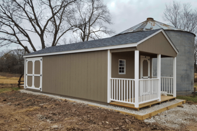 large gable utility shed in ia - beige painted wood siding with white trimmed double doors on long side - white fendced porch with matching door and window on short side all beneath black shingled roof - shed sits besited gray metal silo near leafless trees on earthy property