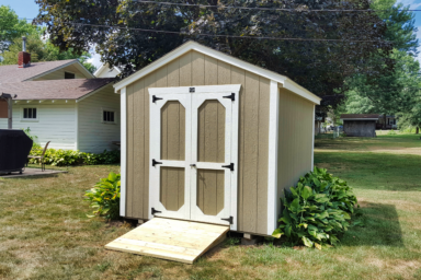 small utility shed in ia - Beige painted wood siding and white -trimmed double doors and corners beneath dark shingled roof - shed is bordered by green vegetation on grassy property with nearby houses and trees