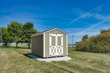 utility shed near me - Beige siding with white-trimmed double doors beneath brown shingled roof - shed sits gravel foundation surrounded by green grass vegetation and a few nearby trees beneath blue sky