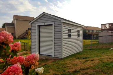 Light gray and white utility shed for sale with two side windows and a white rollup corrugated door beneath dar shingled roof - shed sits in black fenced backyard near vibrant pink flowers - beige houses nearby