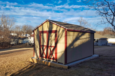 wood utility shed ia - Tan siding and burgundy-trimmed double doors and corners beneath brown shingled roof - shed sits on gravel foundations with several one-story houses and barren trees within view