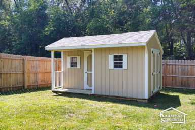 Beautiful backyard cottage shed - beige wood siding with white-trimmed double doors on side - shoite single door overlooking porch and two windows with white shutters on long side - brown shingled roof - shed sits in corner of fenced back yard