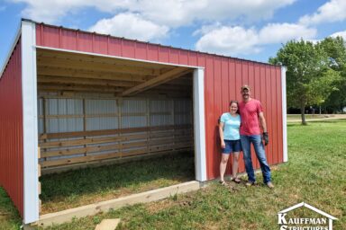 red painted metal loafing shed with white trim around entrance - smiling man and woman standing on grass beside entrance