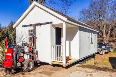 White shed being transported by a shed mover using specialized hydraulic equipment.