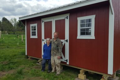 Backyard shed - red wood siding with white-trimmed corners and double doors - smiling older couple stand in front